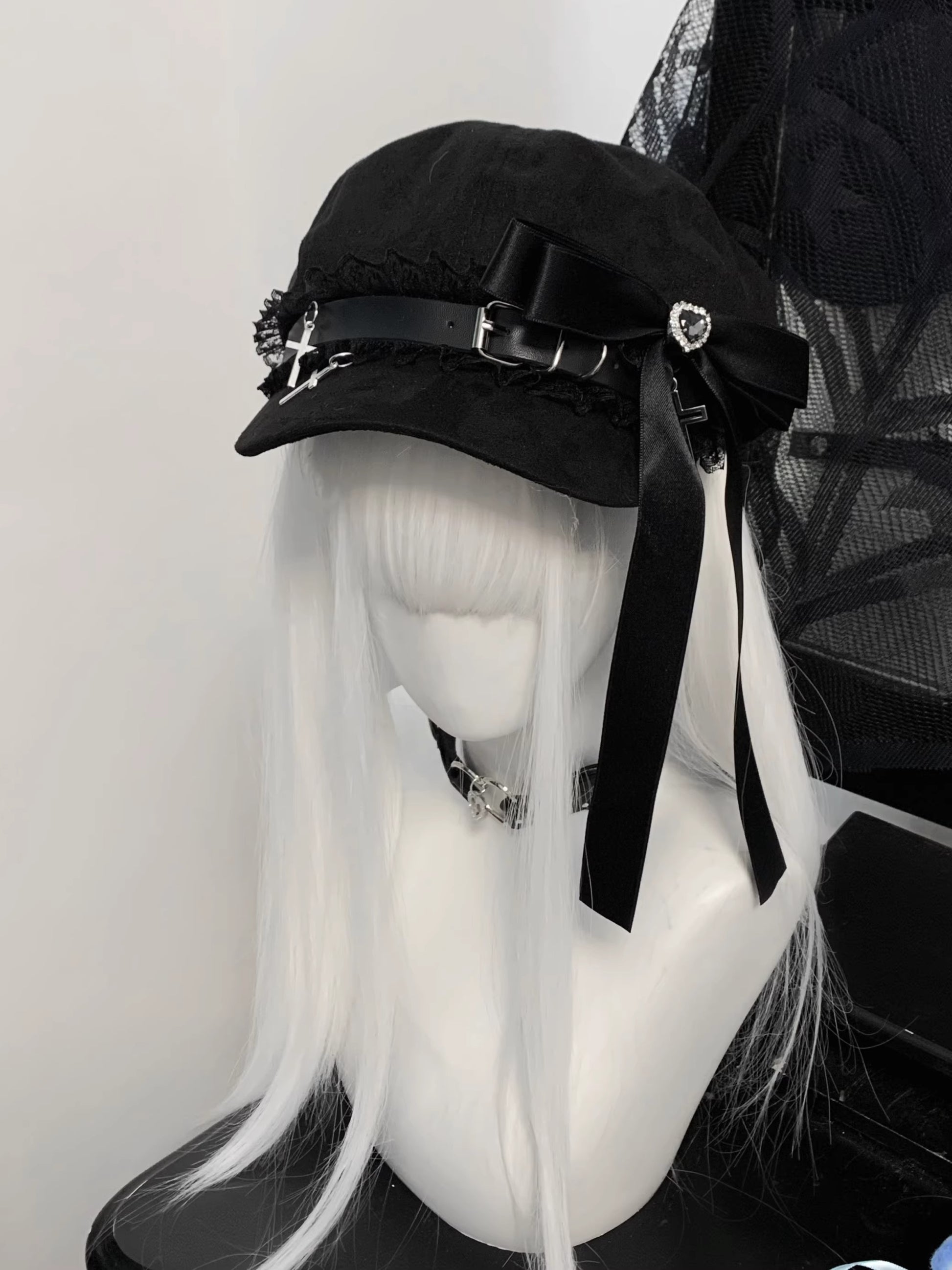 Black Jirai Kei newsboy cap on a white-haired mannequin, featuring a large black satin ribbon, lace trim, and silver cross charms.
