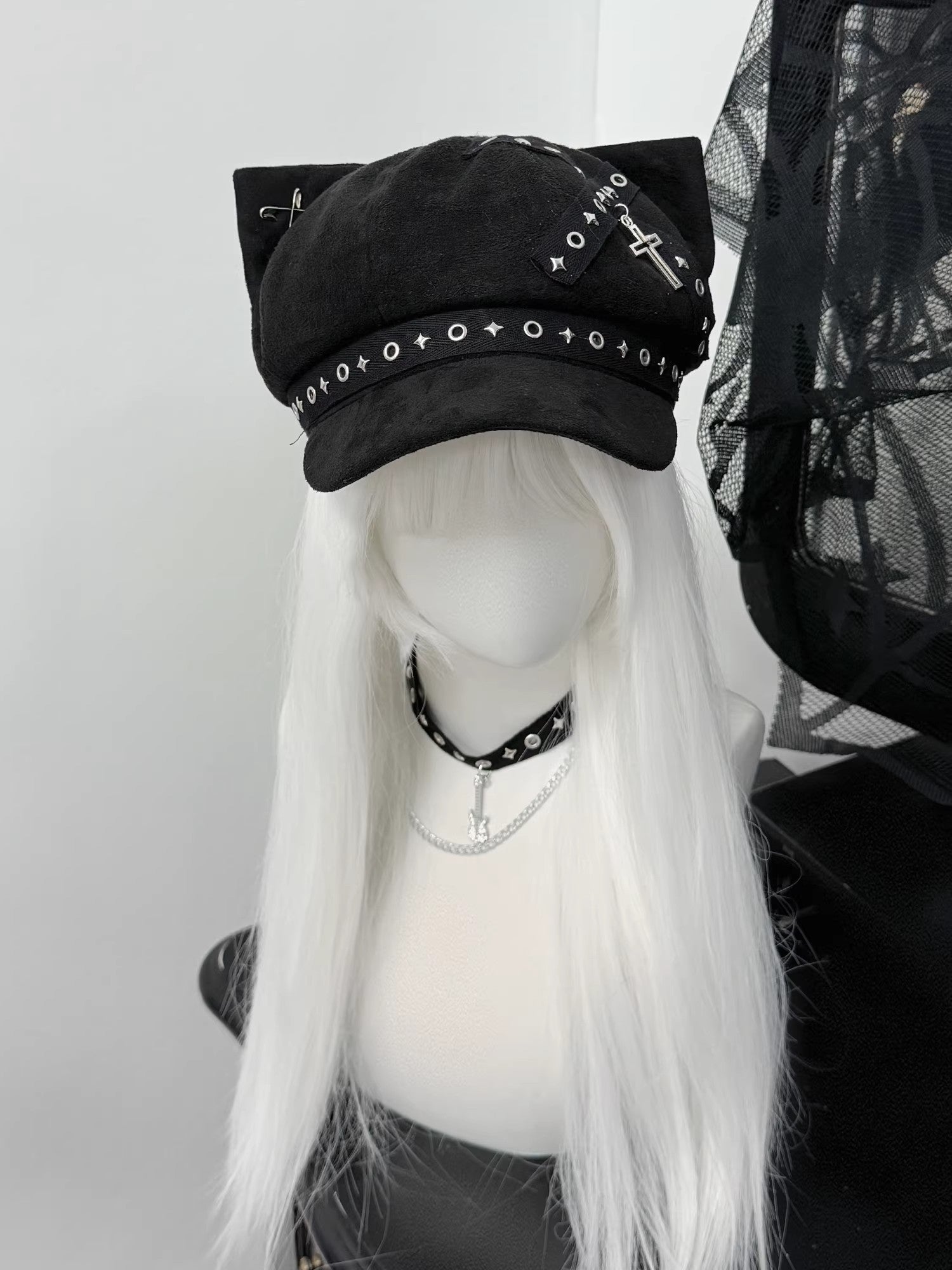 Black cat ear newsboy cap with metal cross charms, safety pins, and grommet webbing on a white-haired mannequin, gothic alt style.