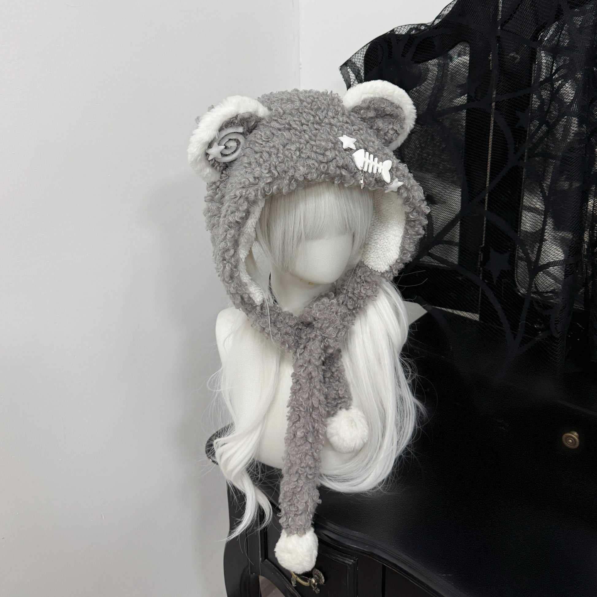 Gray fuzzy bear ear balaclava hat on a white mannequin, featuring white pom-pom ties, a metal fishbone charm, and a spiral patch, Jirai Kei fashion style.