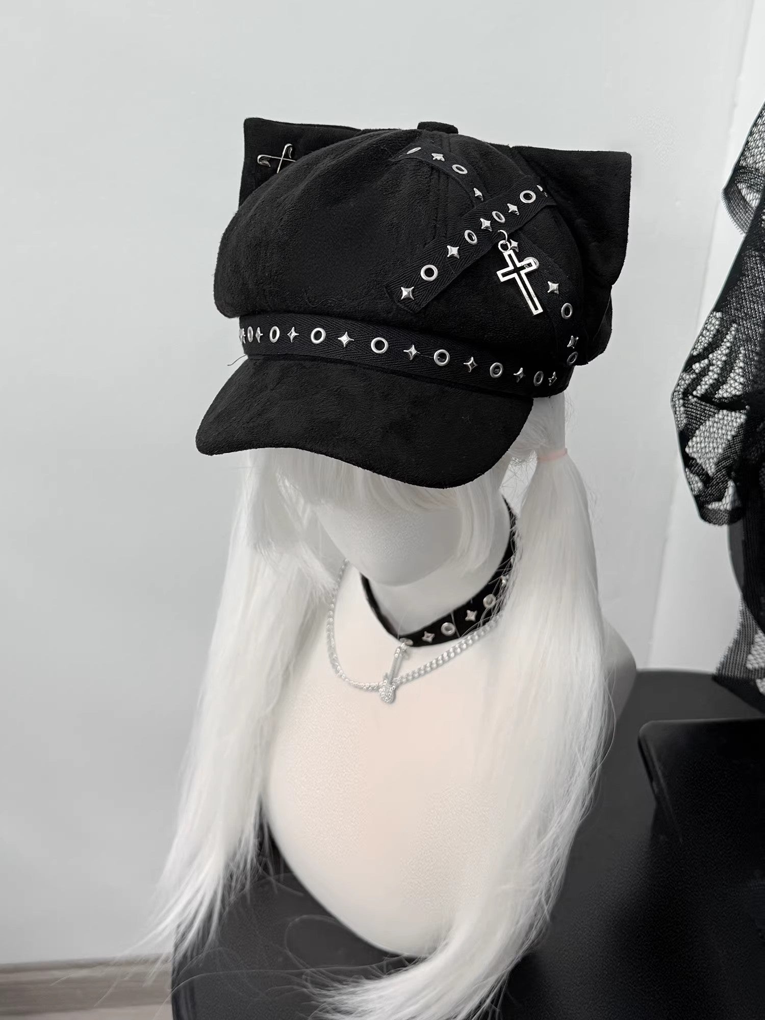 Black cat ear newsboy cap with metal cross charms, safety pins, and grommet webbing on a white-haired mannequin, gothic alt style.