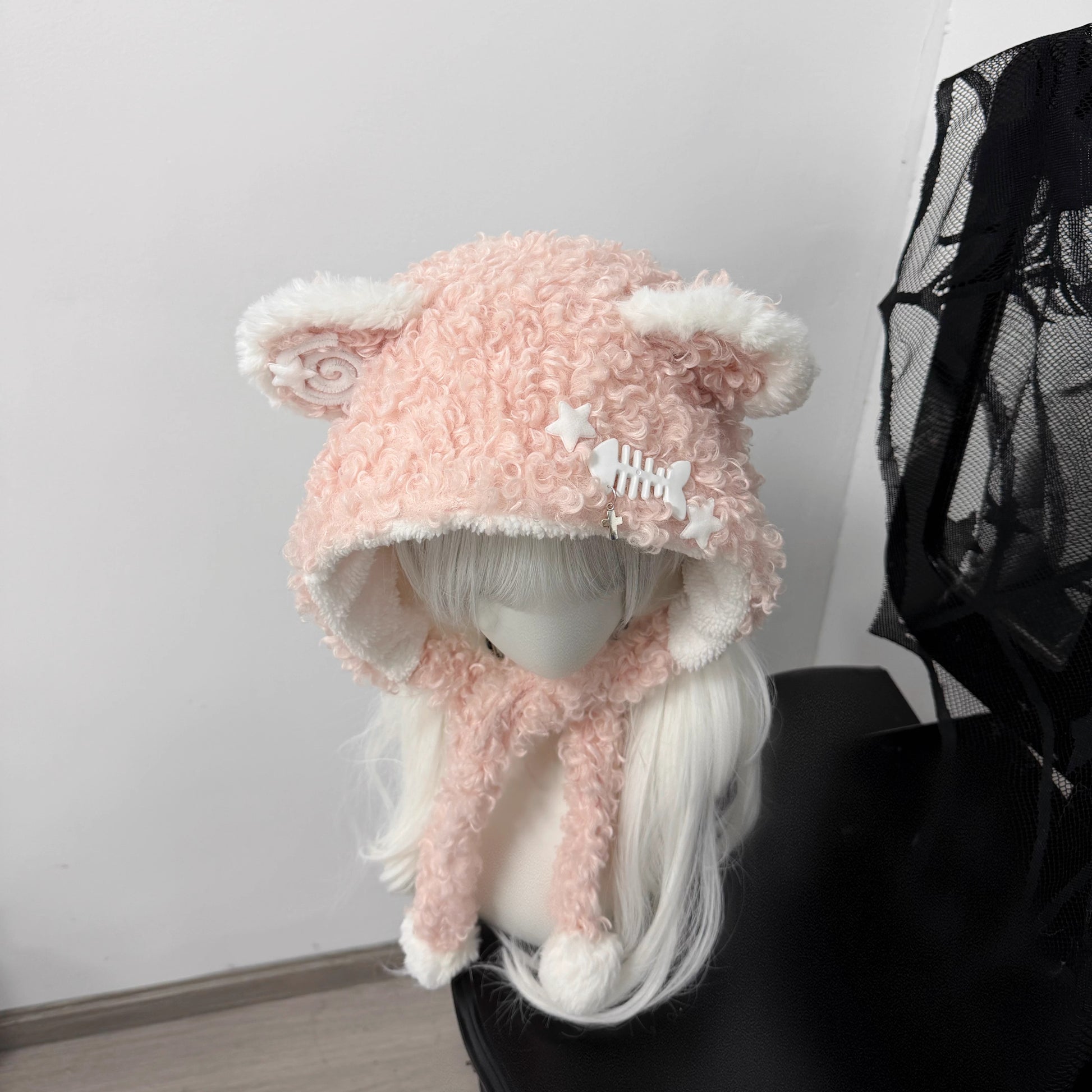 Pink fuzzy bear ear balaclava hat on a white mannequin, featuring white pom-pom ties, a metal fishbone charm, and a spiral patch, Jirai Kei fashion style.